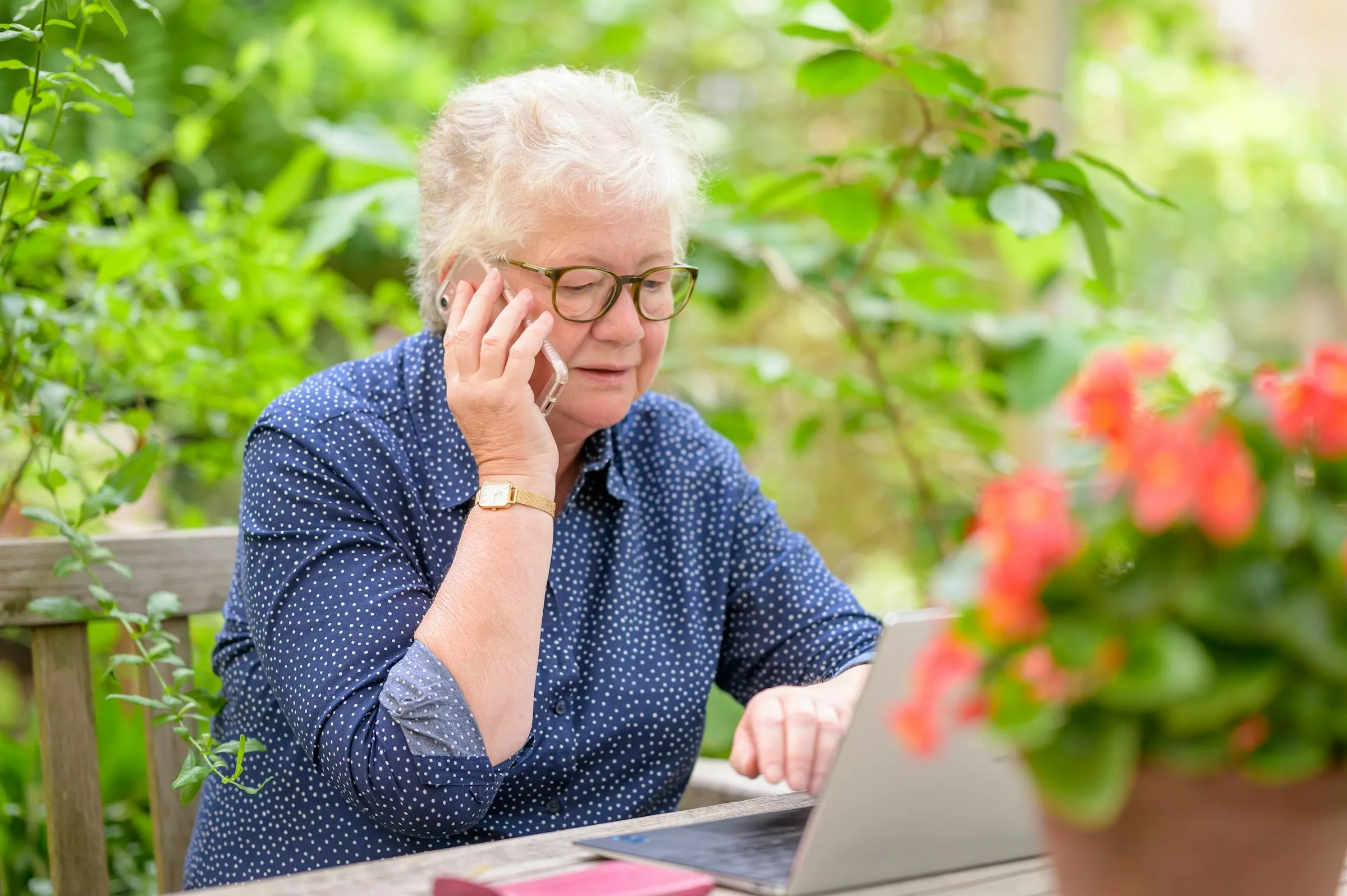 A retiree on the phone reviewing investments on her laptop in a sunny garden